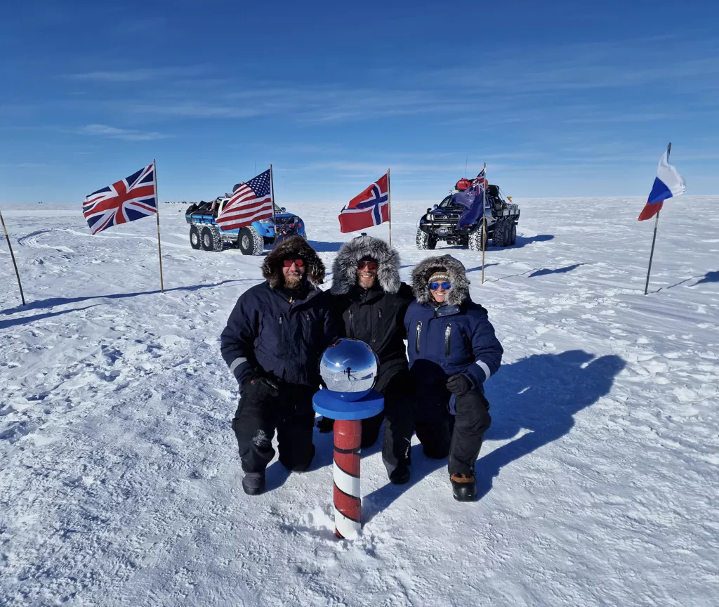 Three people in winter gear kneeling on snow near a striped pole with six-wheeled vehicles and international flags in the background under a clear blue sky.