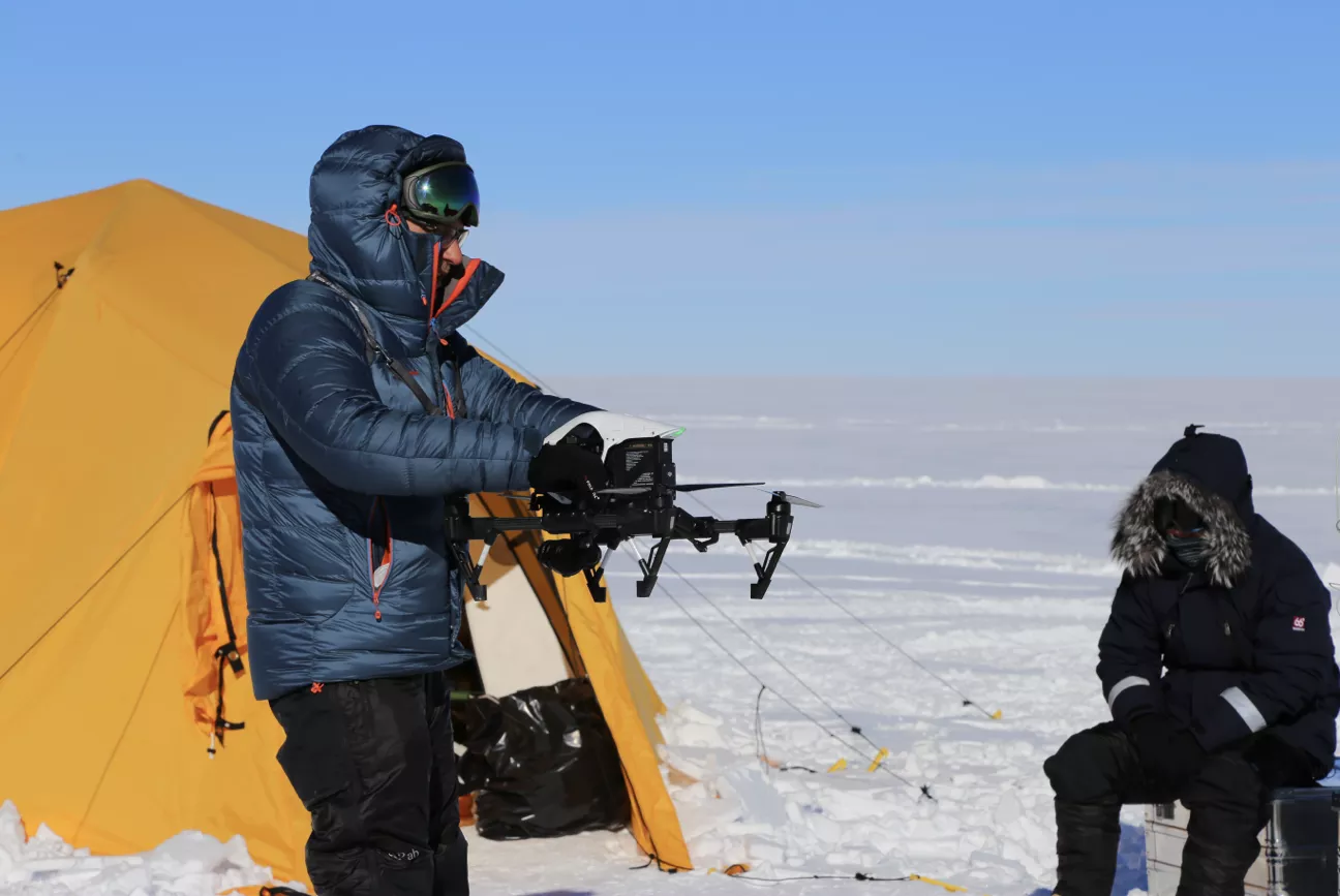 Two people in heavy winter clothing near a yellow tent on snow; one is holding a drone while the other sits on a box.