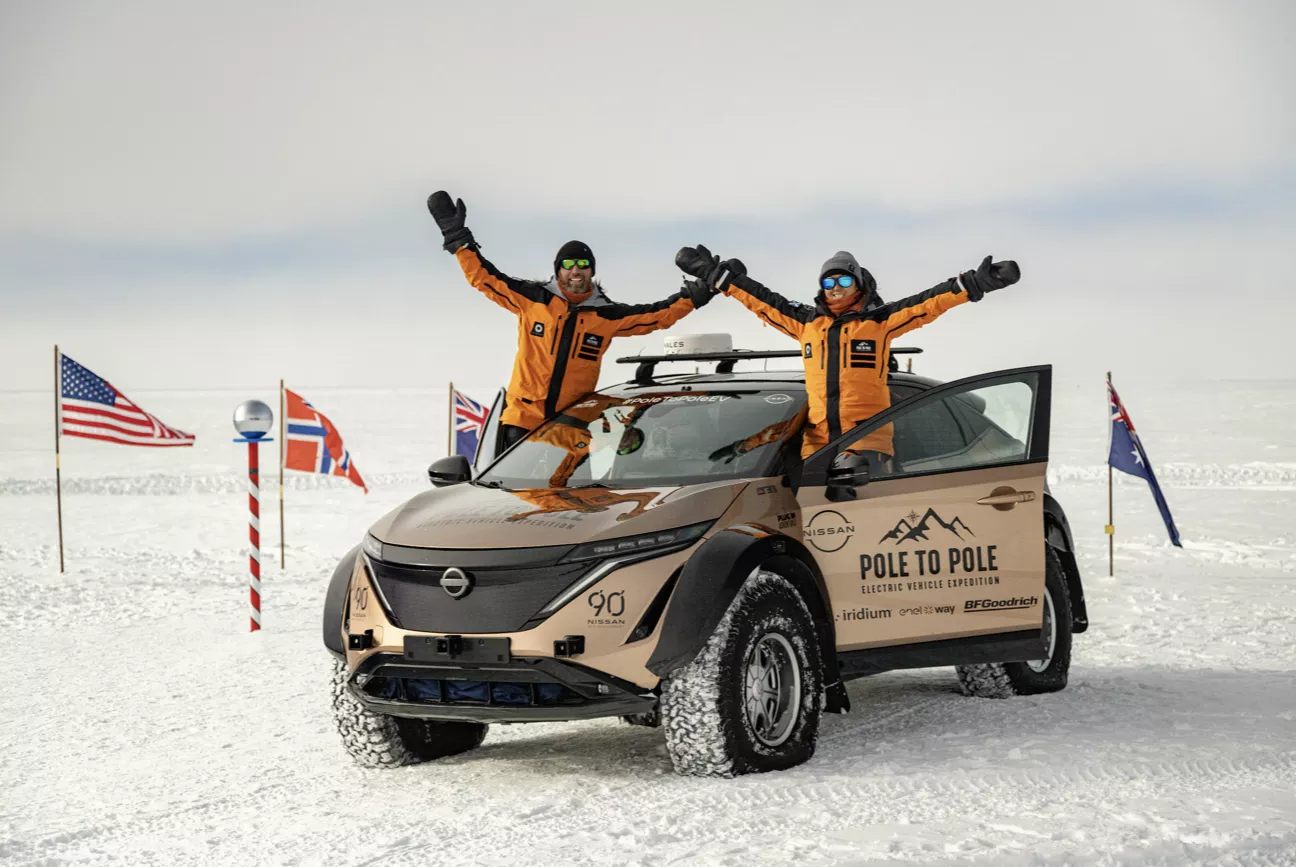 Two people in orange jackets with raised arms standing beside and partly inside a beige Nissan electric vehicle with 'Pole to Pole Electric Vehicle Expedition' branding on snowy terrain surrounded by several international flags.
