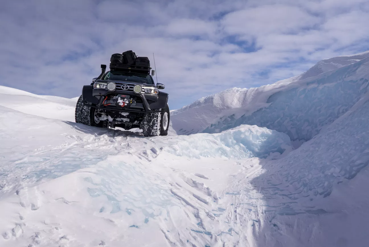 Toyota off-road vehicle equipped with large tires and roof luggage driving on a snowy, icy landscape under a cloudy sky.