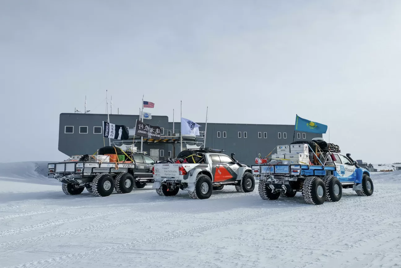 Three off-road trucks with large tires and cargo on snowy ground in front of a large gray building with flags, including the American and Kazakhstan flags.