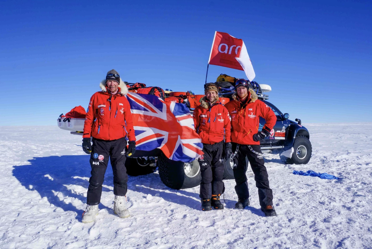 Three explorers in red jackets standing on snow in front of a vehicle with a Union Jack flag and a red flag under clear blue sky.