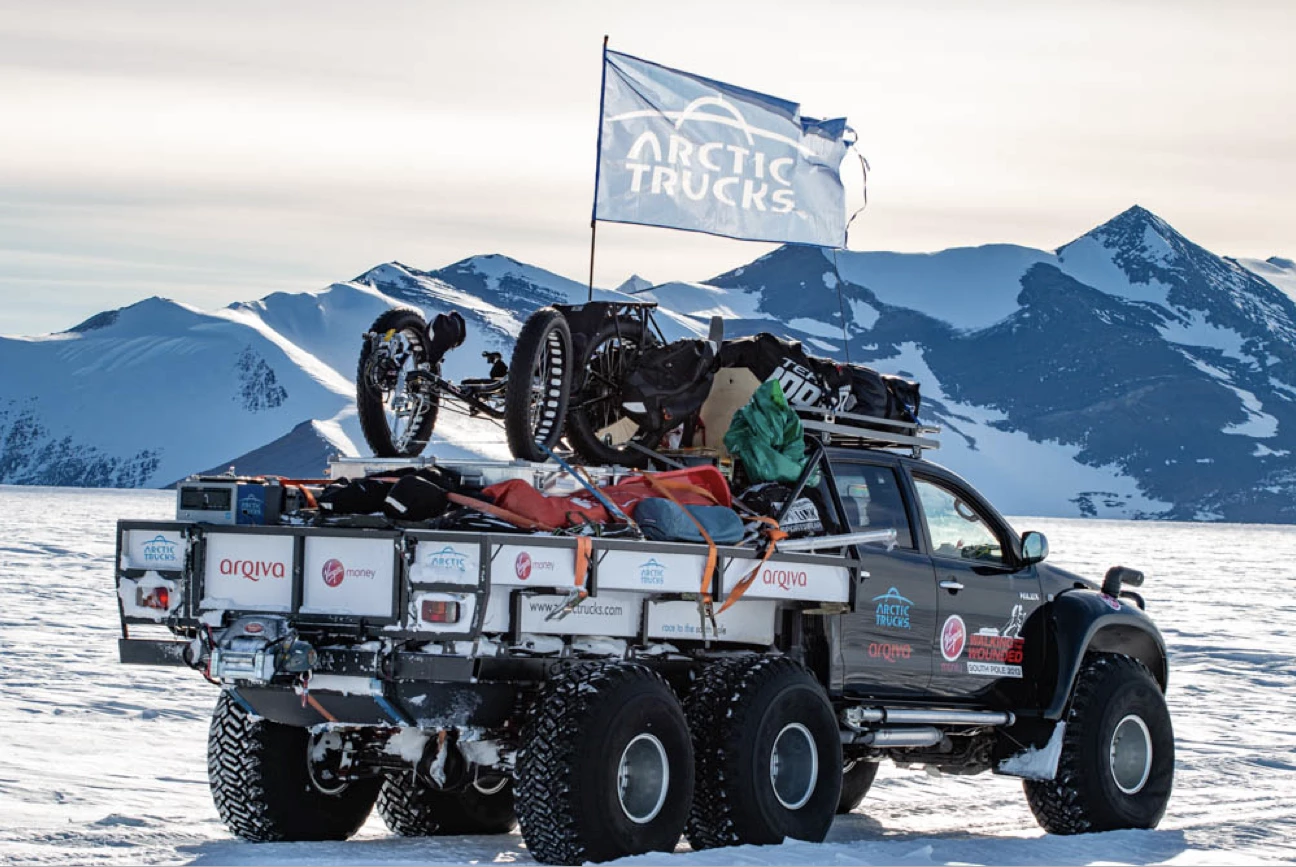 Six-wheeled Arctic Trucks expedition vehicle loaded with gear and a large flag on a snowy landscape with mountains in the background.