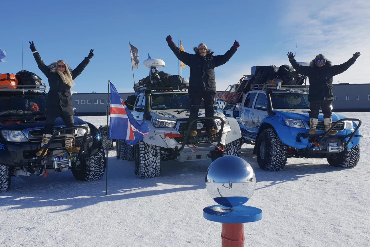 Three people in winter gear standing on off-road vehicles with raised arms on a snowy landscape under a clear blue sky.