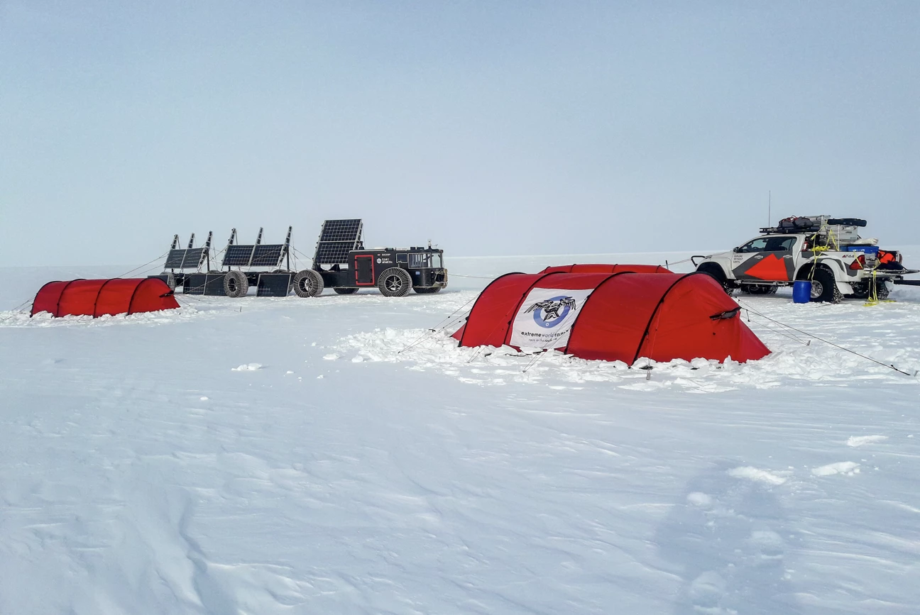 Red expedition tents set up on a snowy plain with a solar panel-equipped vehicle and a white pickup truck loaded with gear nearby.