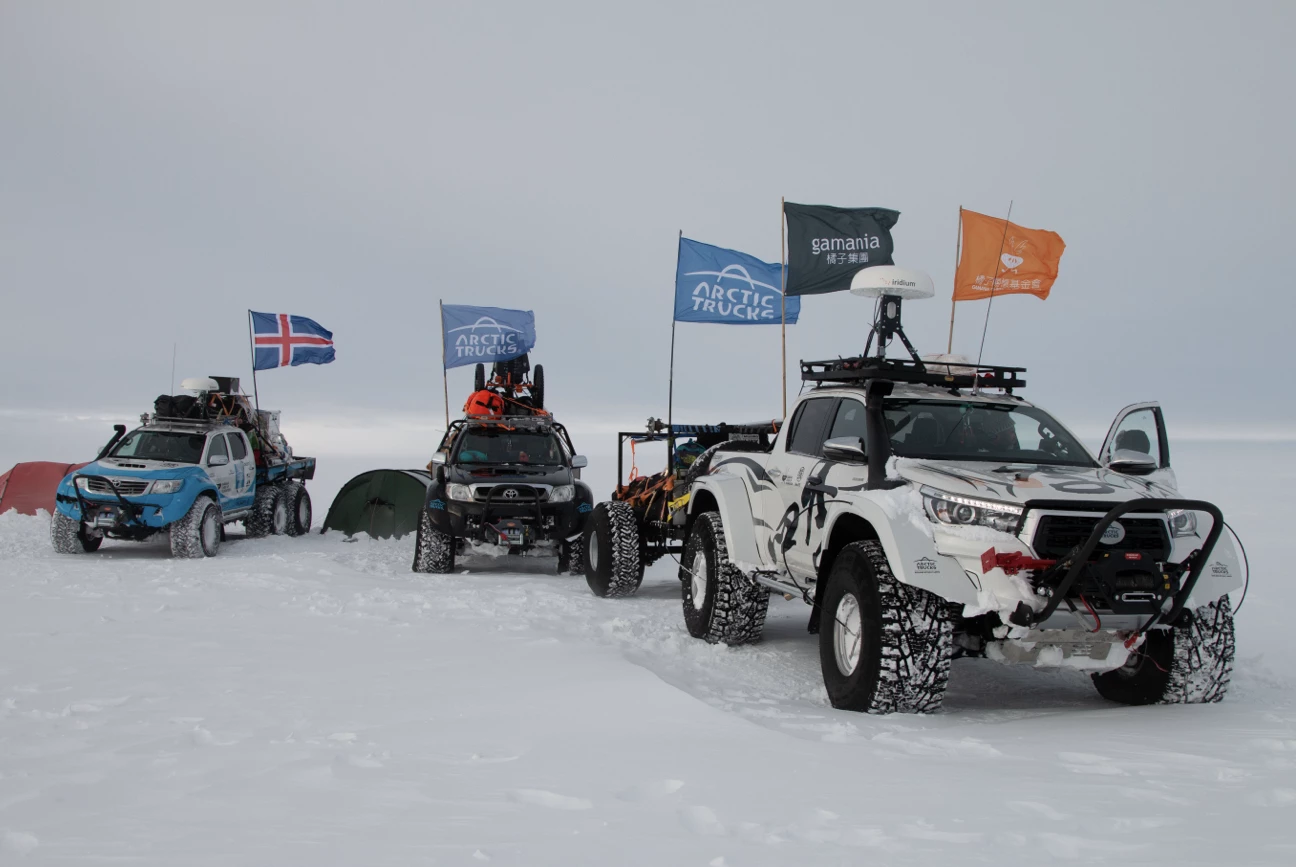 Three off-road trucks with flags parked on snowy terrain beside tents under a gray sky.