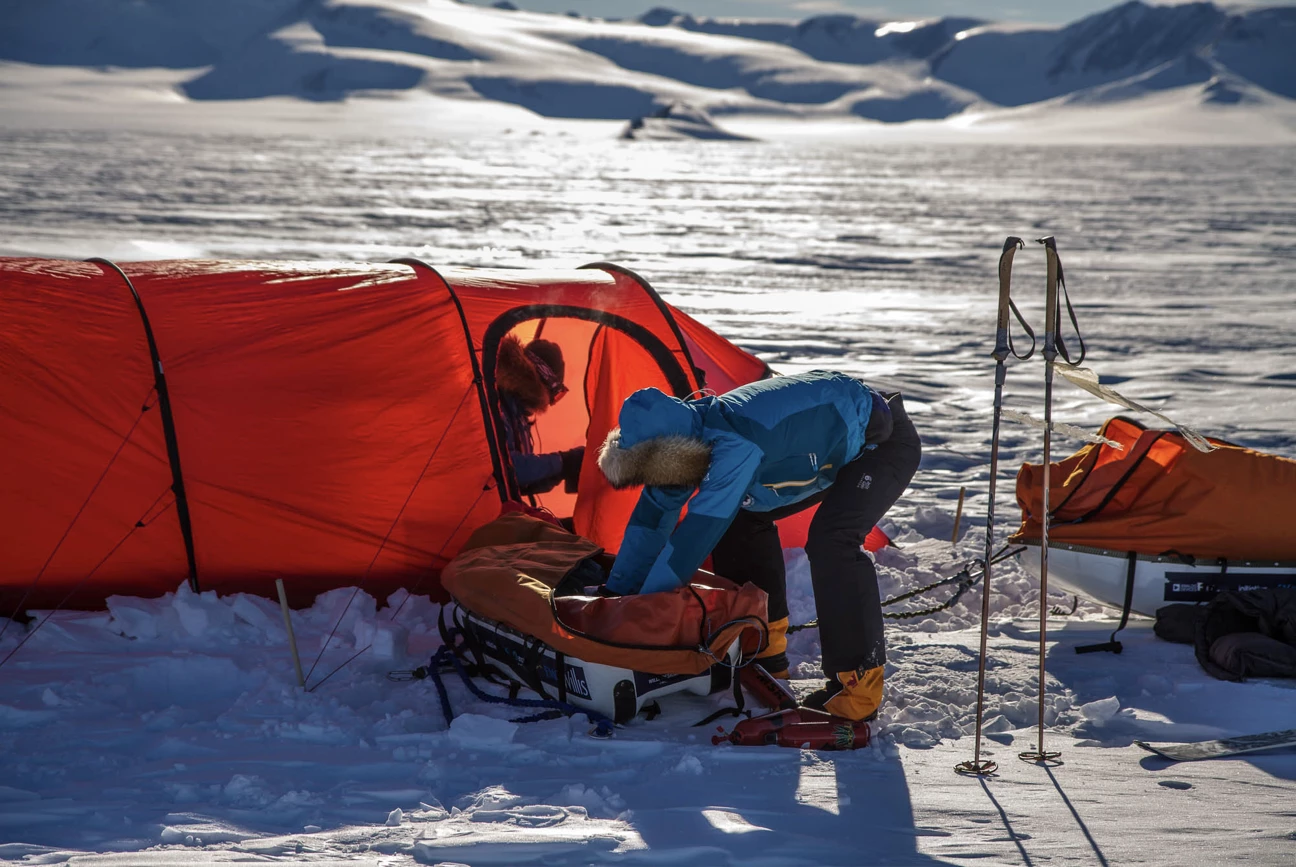 Person in blue winter gear packing equipment next to a red tent on snowy terrain with ski poles standing nearby.