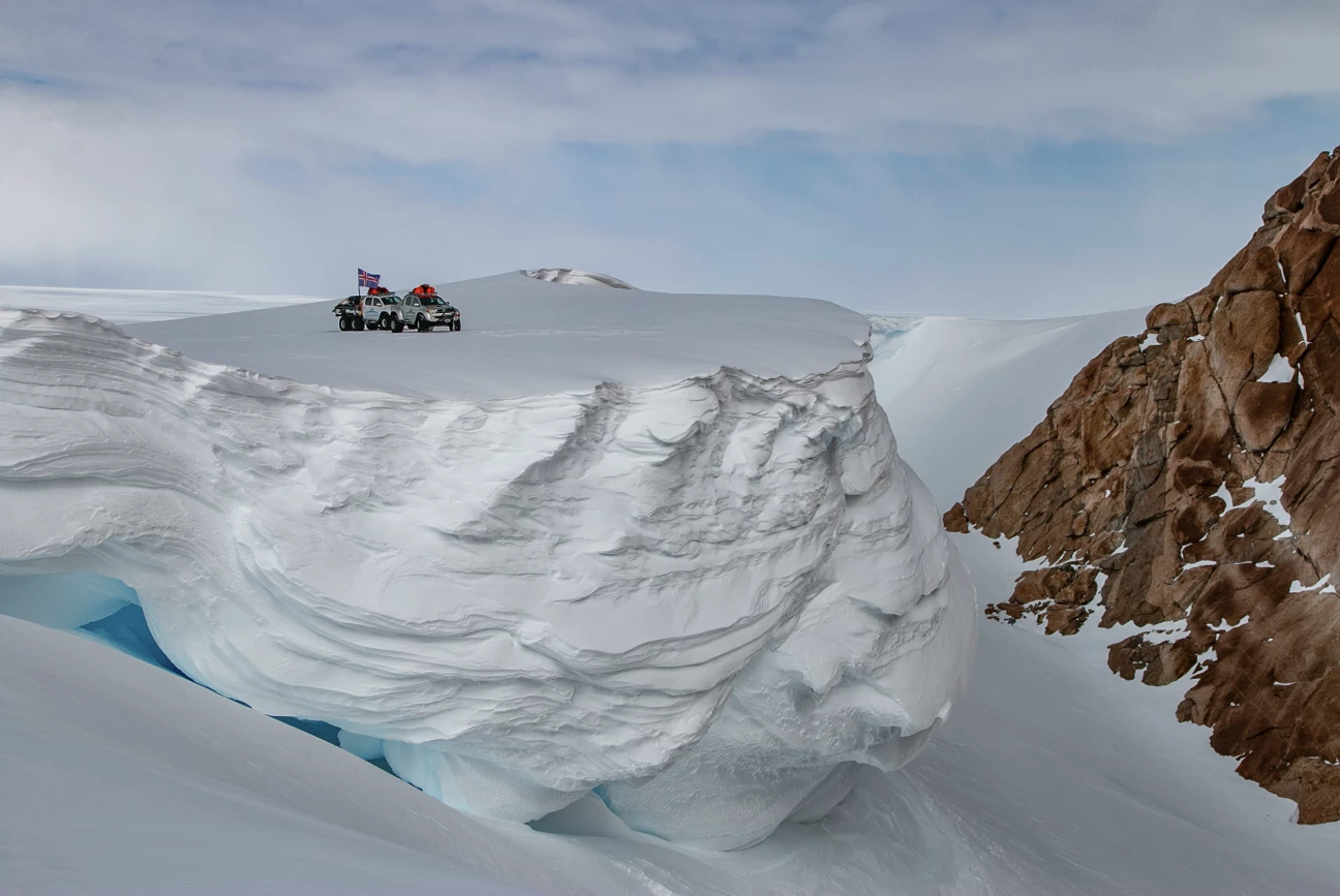 Two vehicles with an Icelandic flag parked on a large snow-covered ice formation near rocky terrain under a cloudy sky.