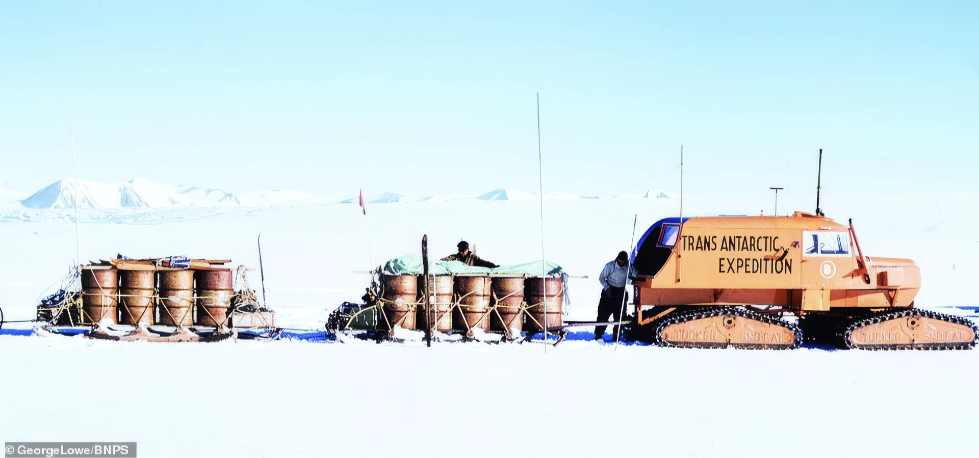 Orange tracked vehicle labeled 'Trans Antarctic Expedition' towing sleds loaded with barrels across a snowy Antarctic landscape with two people managing the equipment.