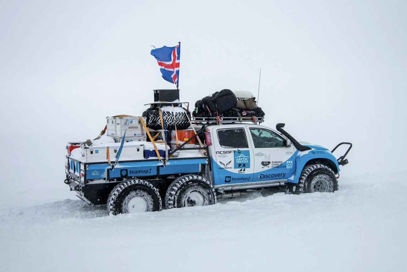 Six-wheeled blue and white Arctic expedition truck with gear and Icelandic flag on snowy terrain.