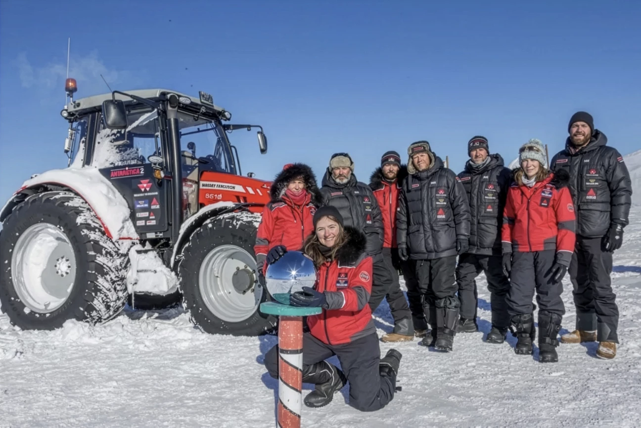 Group of eight people dressed in winter gear standing on snow beside a red Massey Ferguson tractor under a clear blue sky, with one woman kneeling and touching a reflective spherical marker.