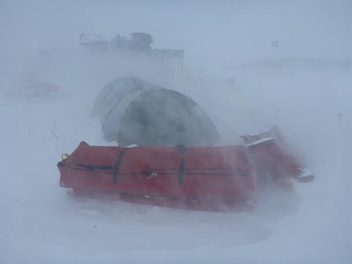 Snow-covered tents and gear partially obscured by blowing snow in a harsh winter environment.
