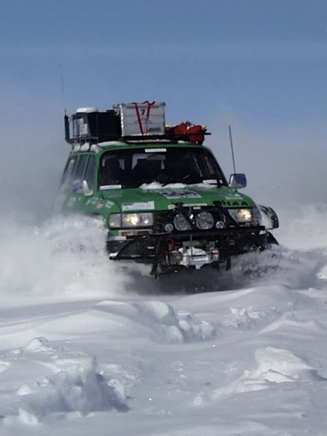 Green off-road SUV driving through deep snow with equipment on roof under clear blue sky.