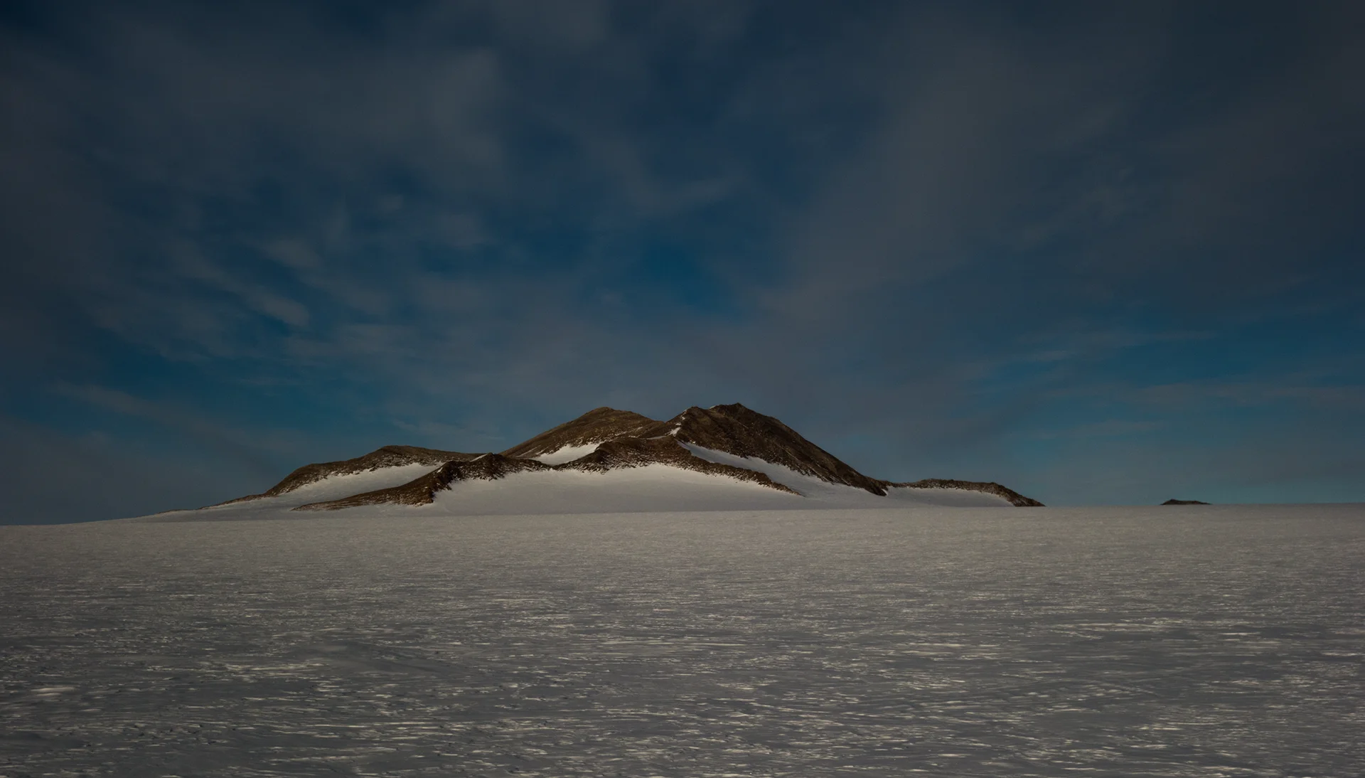 Snow-covered flat terrain with a dark rocky mountain ridge under a cloudy blue sky.