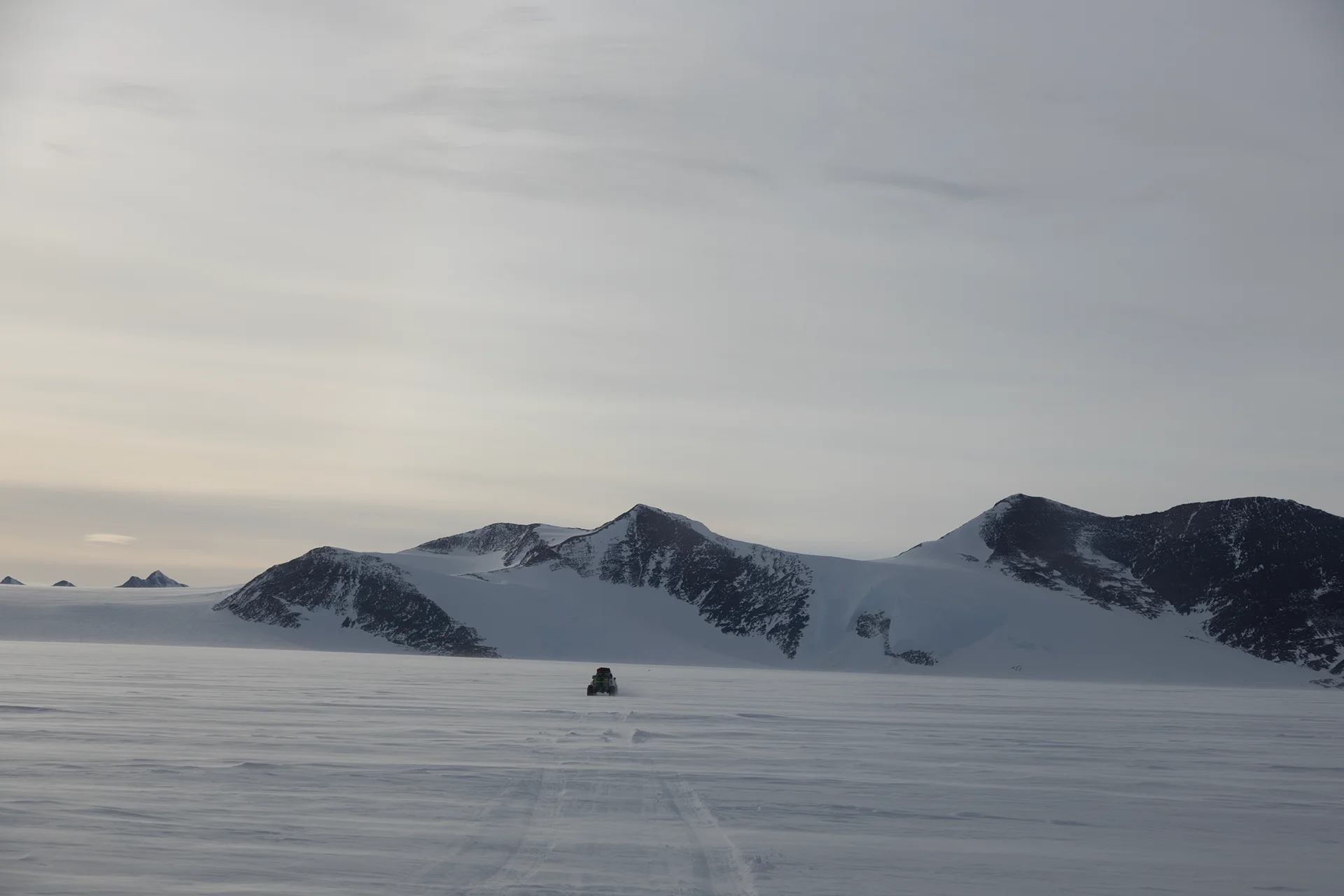 A lone vehicle driving on a vast snowfield toward dark, snow-capped mountains under a cloudy sky.