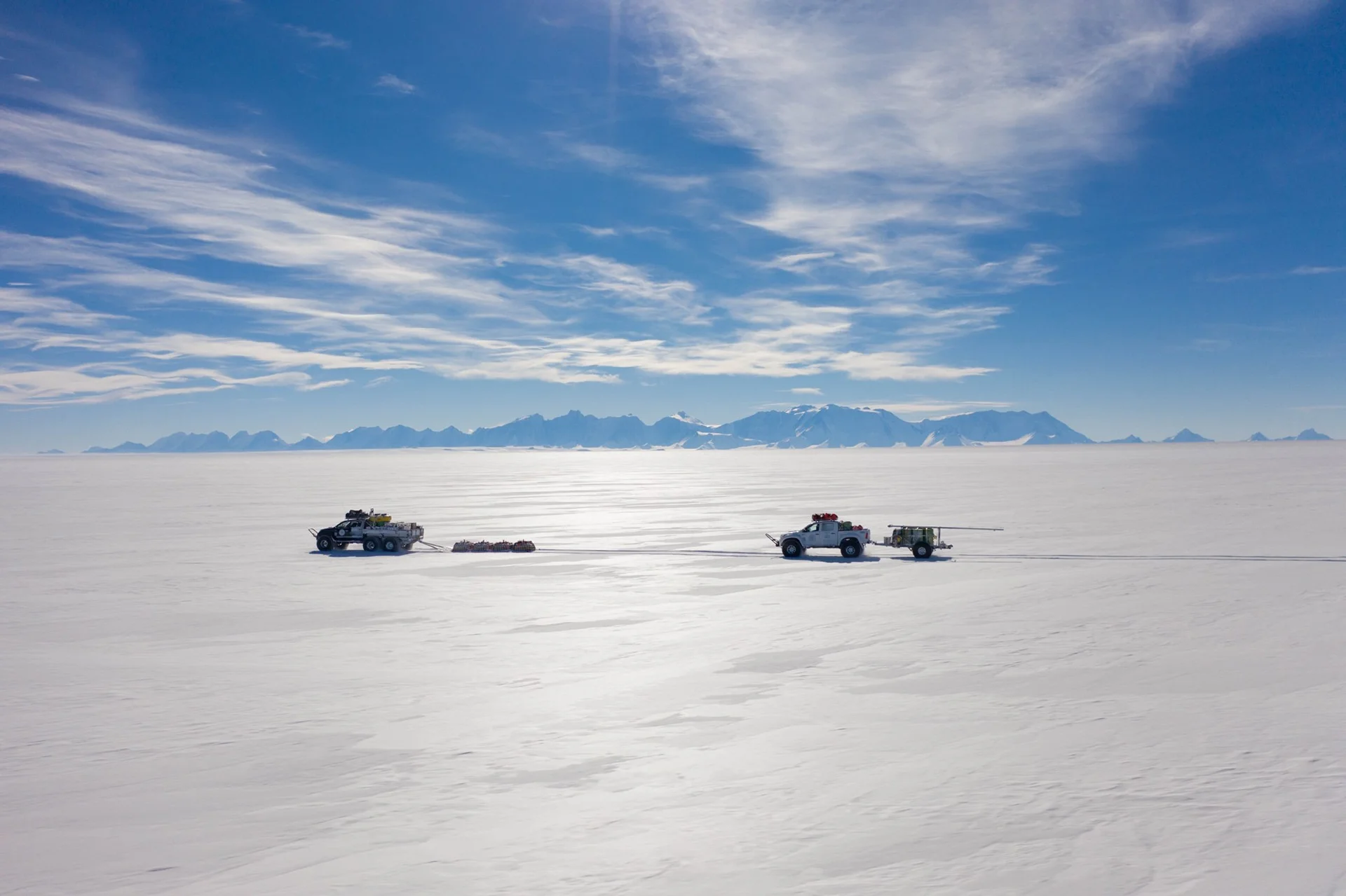 Two vehicles towing sleds across a vast snowy ice field with mountains under blue sky.