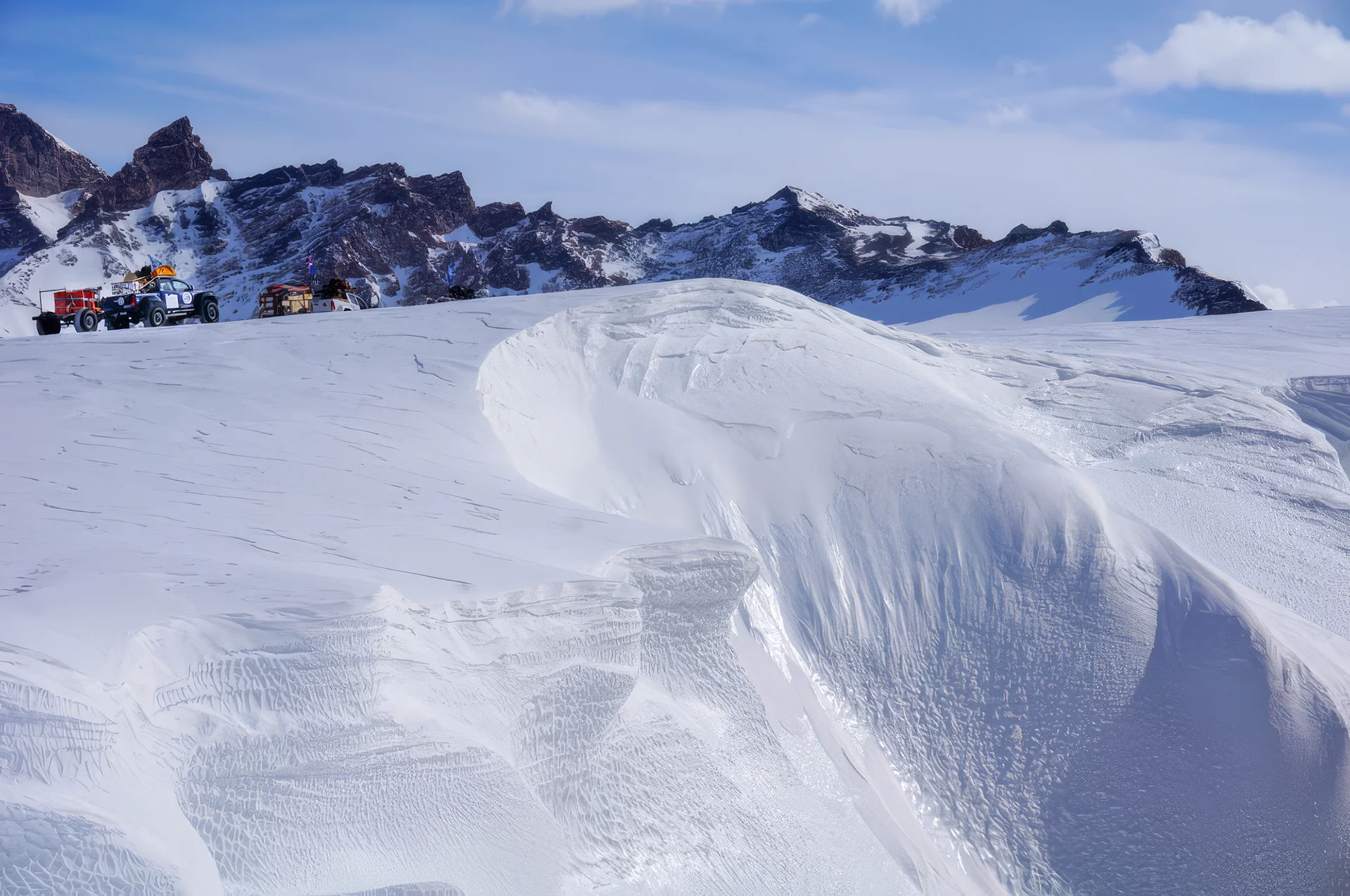 Snow-covered landscape with vehicles and rocky mountain peaks under a partly cloudy sky.