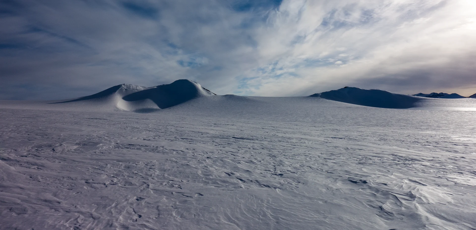 Snow-covered Antarctic landscape with rounded ice hills beneath a cloudy sky.