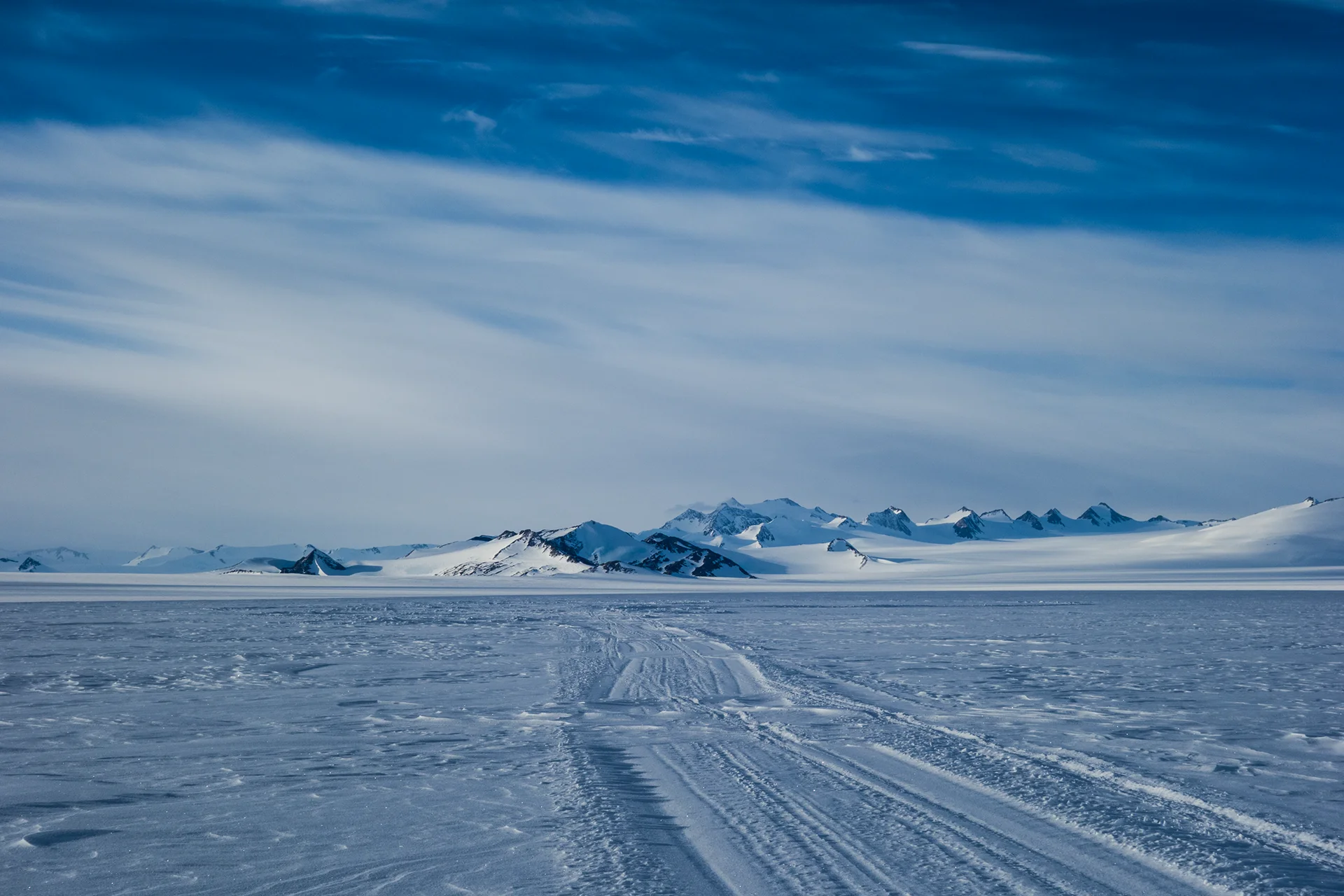 Snow tracks lead across an icy plain toward distant snow-covered mountains under a cloudy blue sky.
