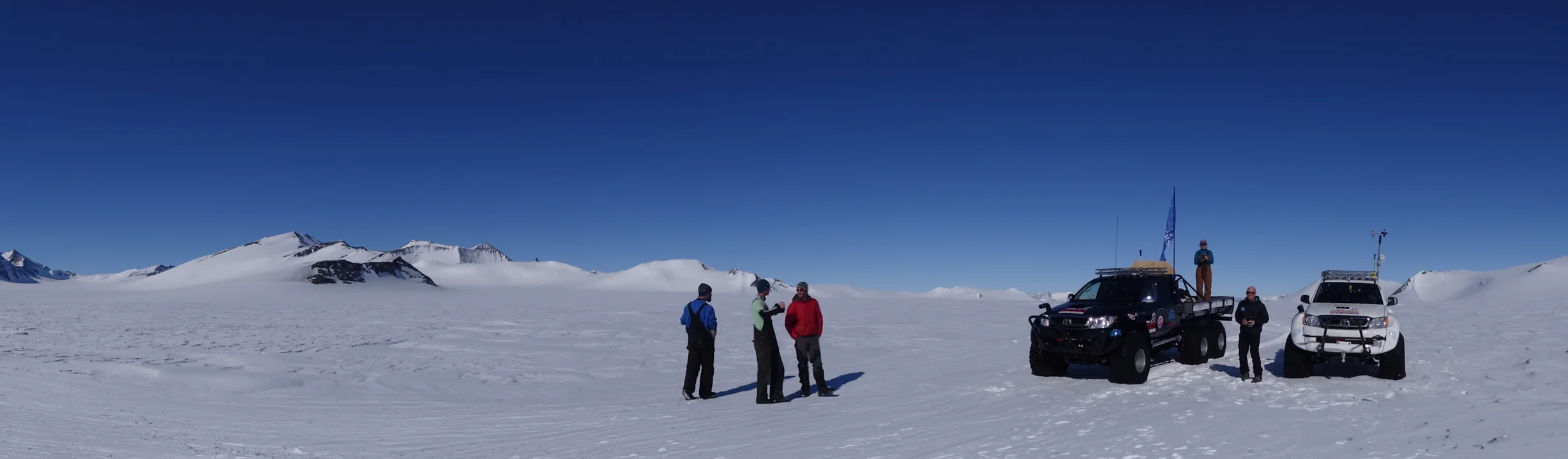Three people standing on snowy terrain near two off-road vehicles under a clear blue sky.