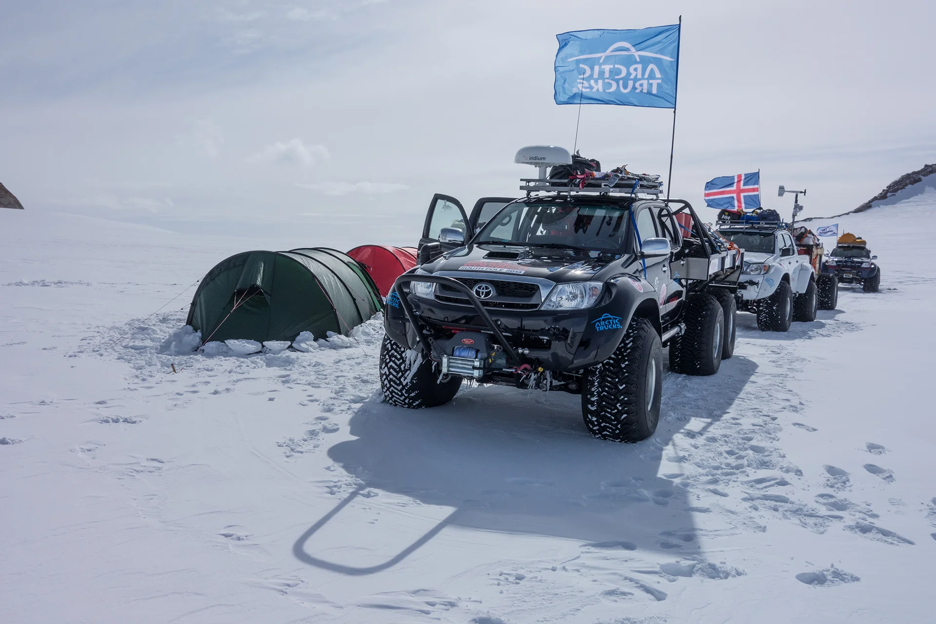 Line of rugged pickup trucks with Arctic Trucks flags parked near tents on snowy terrain.