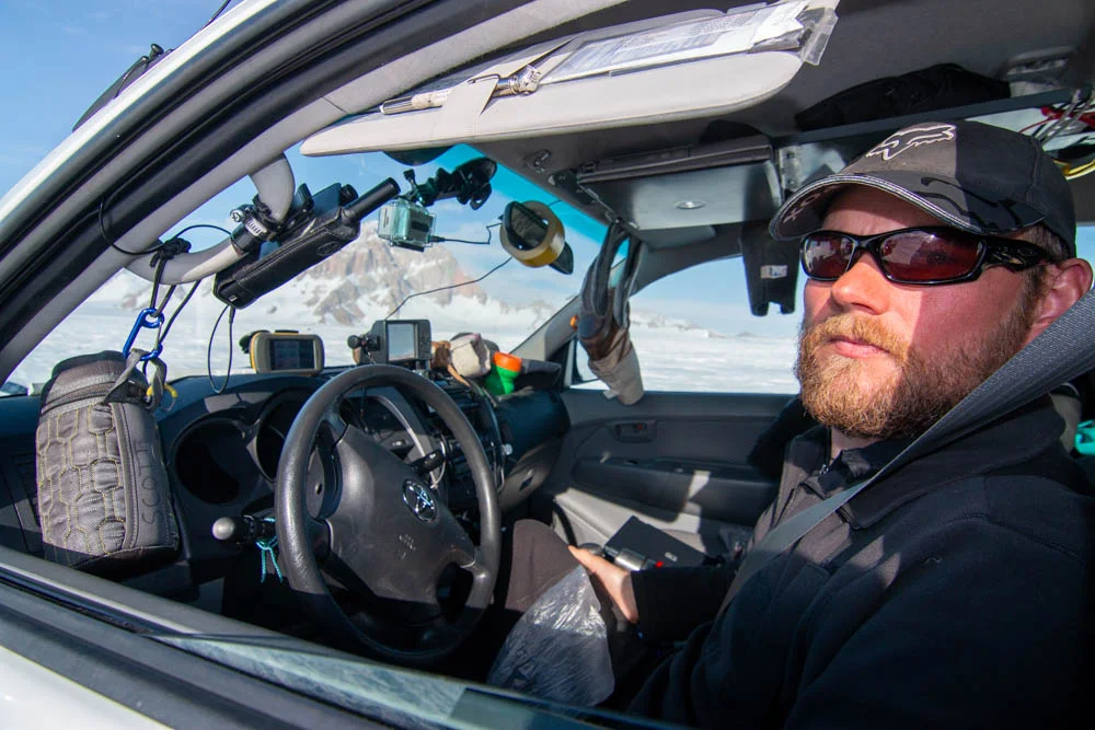 Bearded man wearing sunglasses and cap seated inside a vehicle with snowy mountains outside.