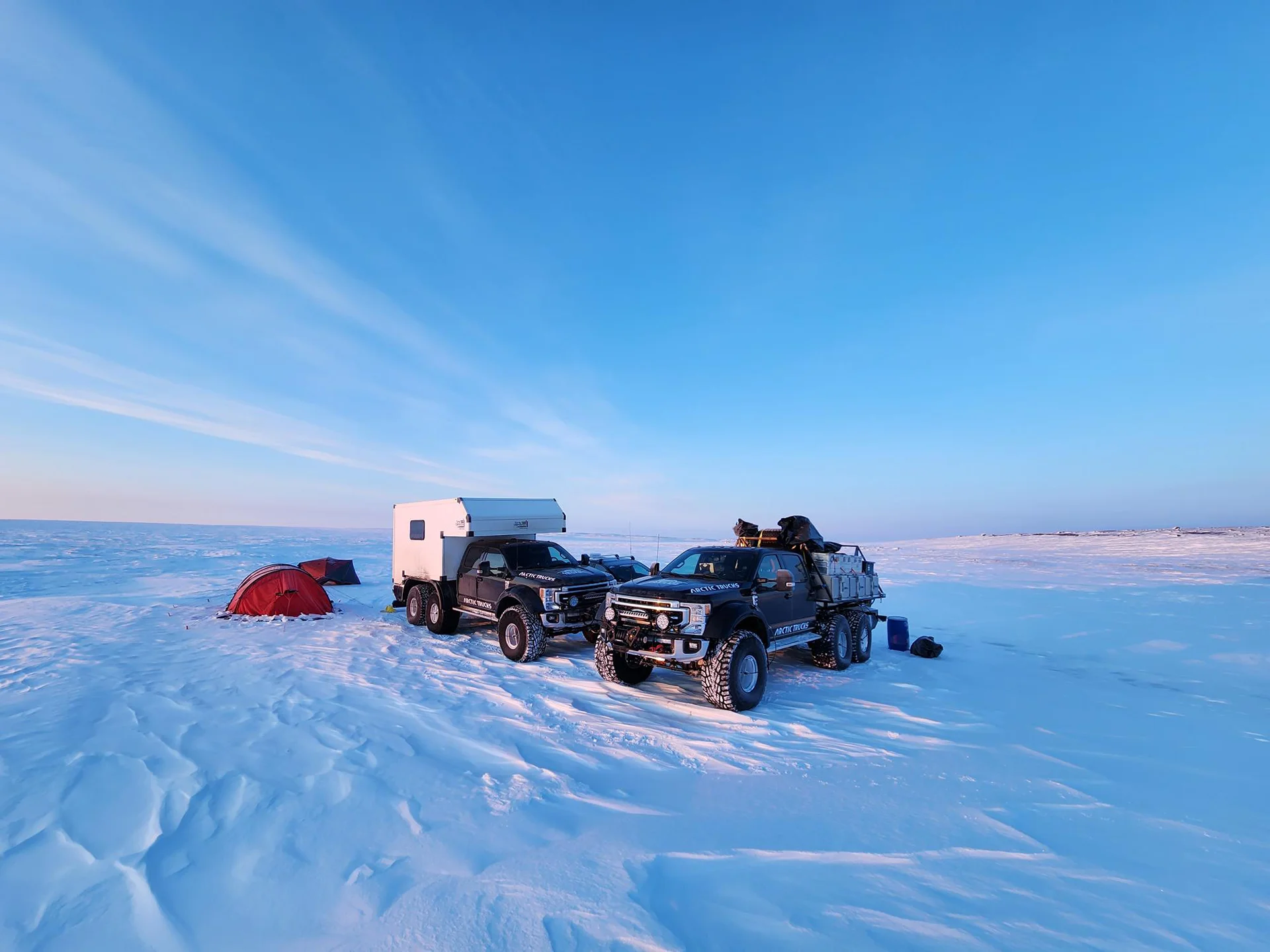 Two six-wheel Arctic Trucks on snow beside two red tents under a clear blue sky at sunset.