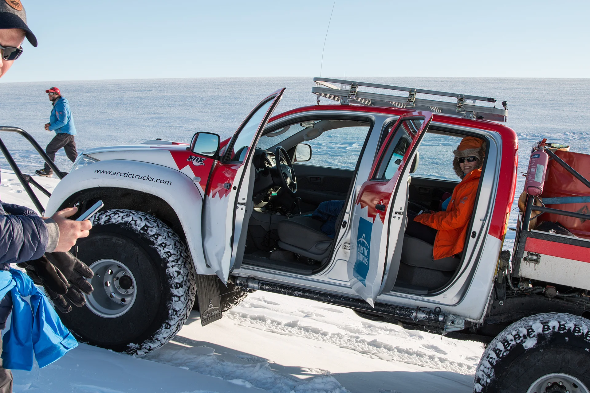 Person in orange jacket sitting in open door Arctic truck on snow with others nearby.