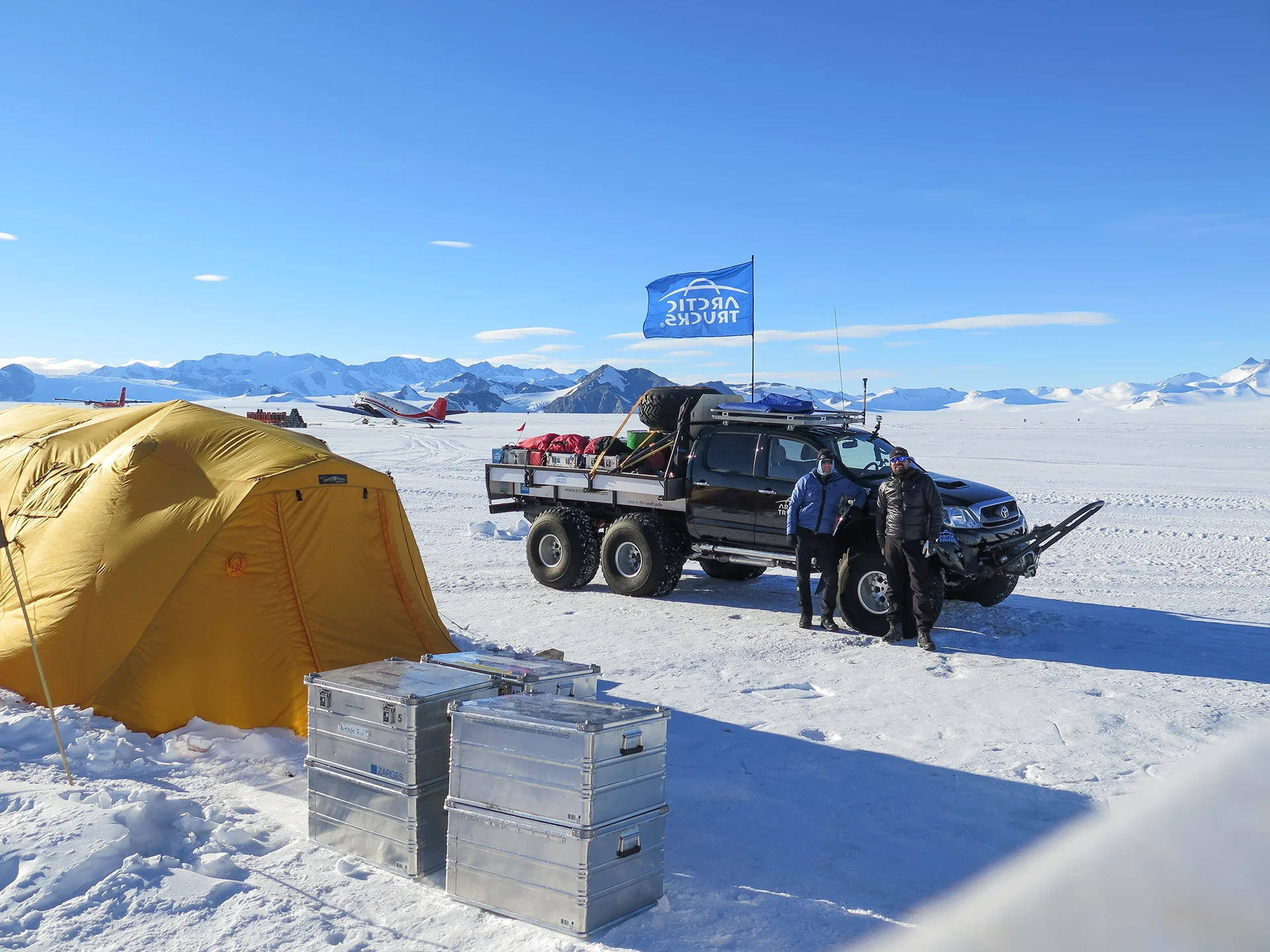 Two people in winter gear stand by a six-wheeled truck and yellow tent on a snowy mountain field.