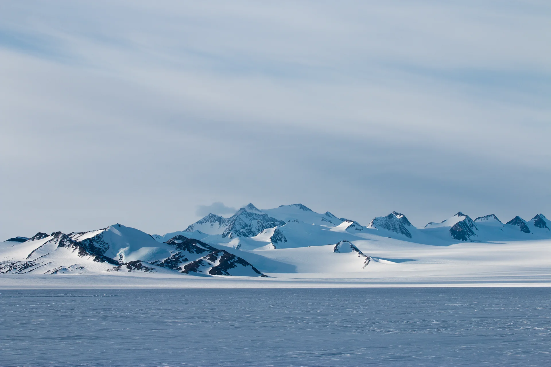 Snow-covered mountain range under a cloudy blue sky with icy plain in the foreground.