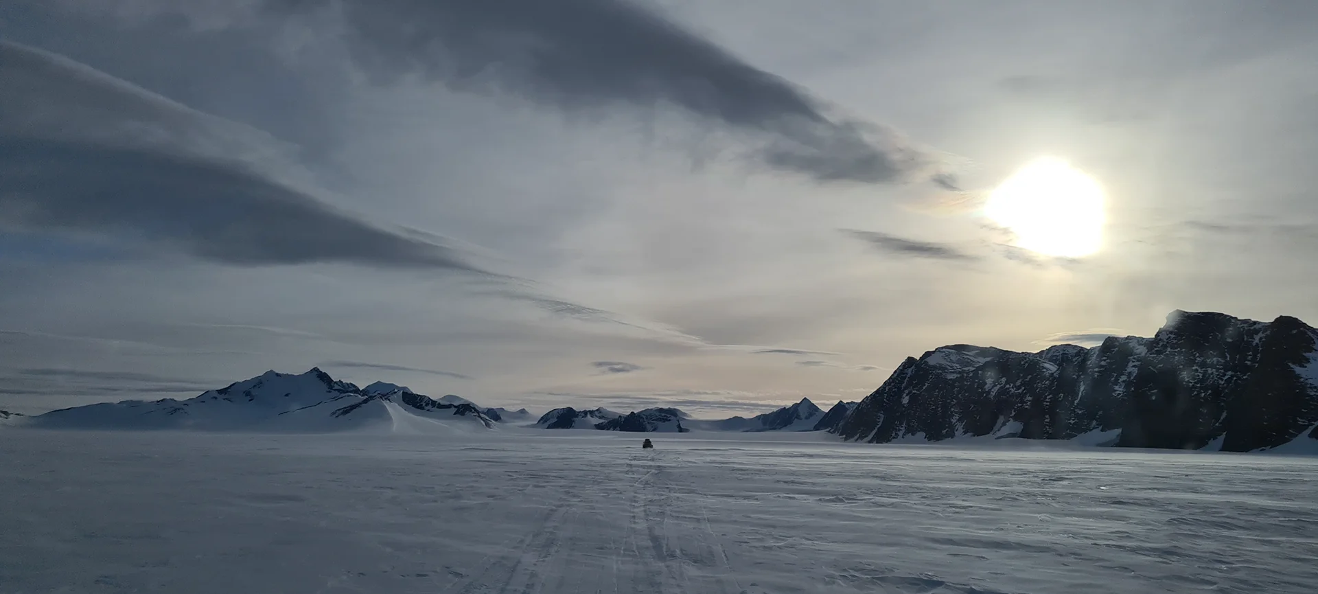 Snowy mountain landscape under a cloudy sky with sun shining and snow vehicle tracks in the snow.