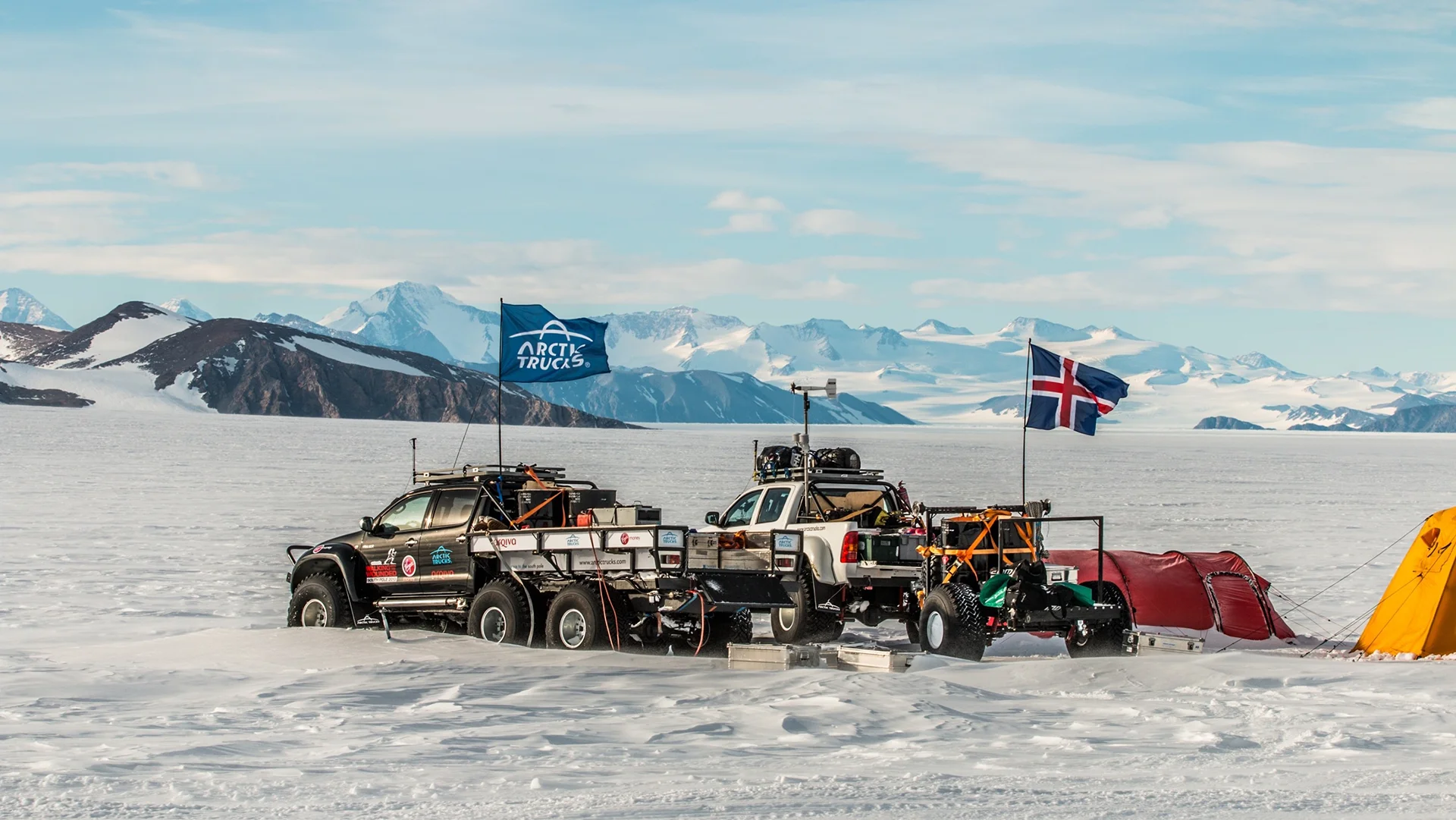Two off-road trucks with trailers and flags parked on snowy terrain with mountains in the background.