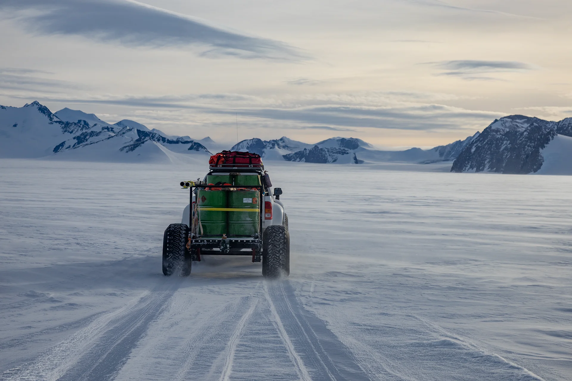 Off-road vehicle carrying barrels and gear driving on snow with mountain range in the background.