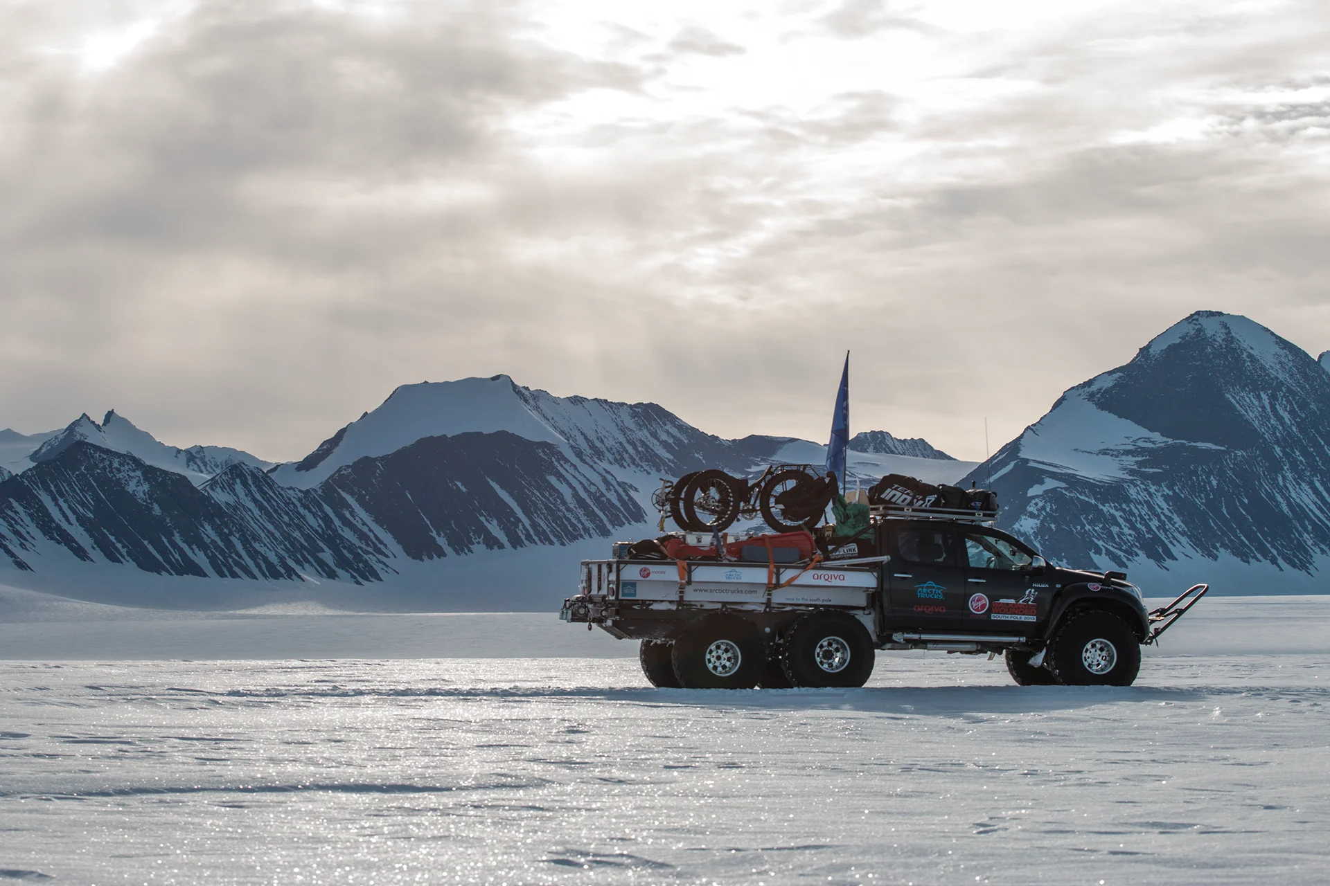 A six-wheeled expedition truck with equipment and a bike crosses a snowy plain with mountains in the background.