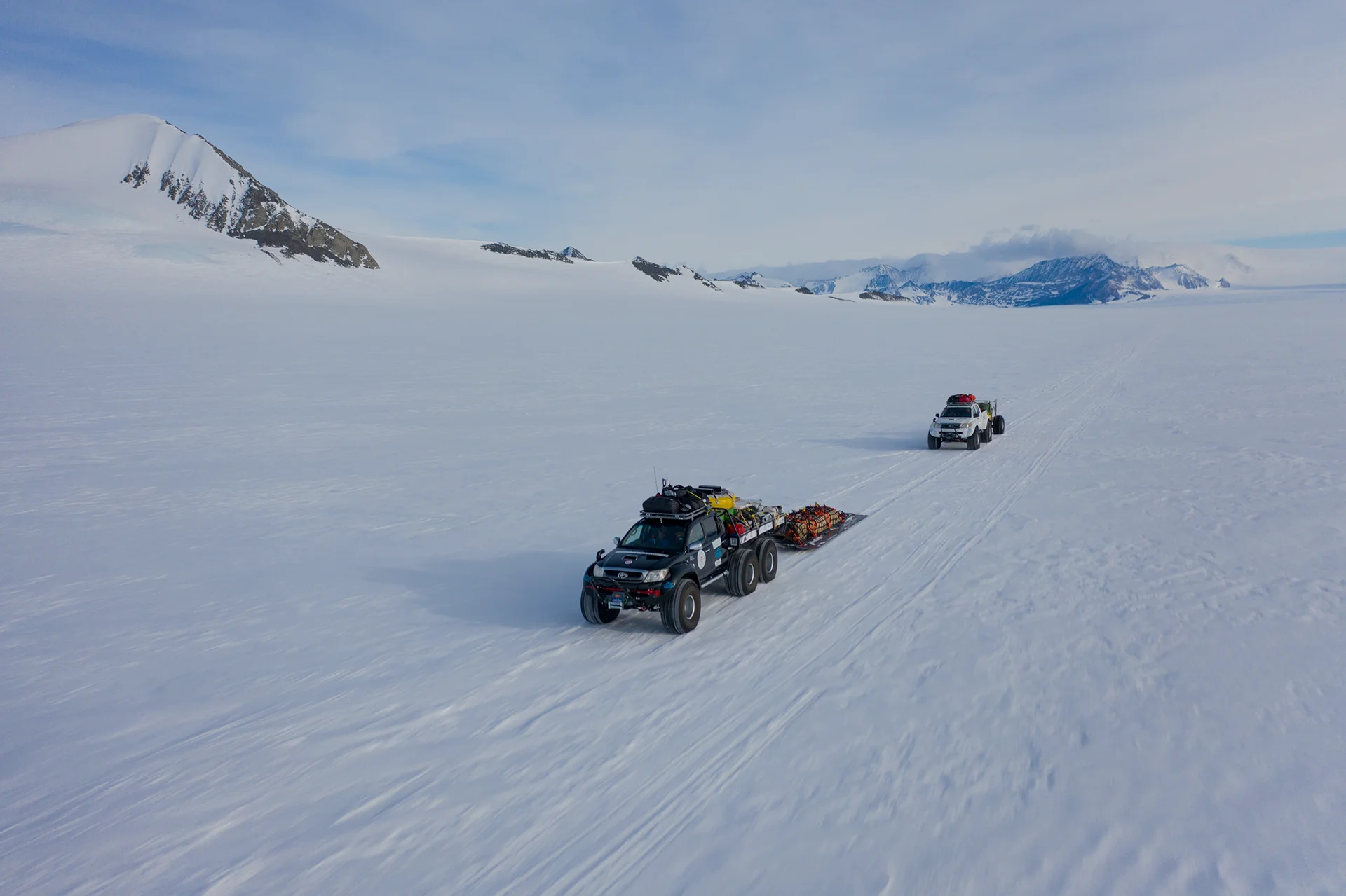 Two six-wheeled vehicles pulling sleds across a snowy plain with mountains in the background.