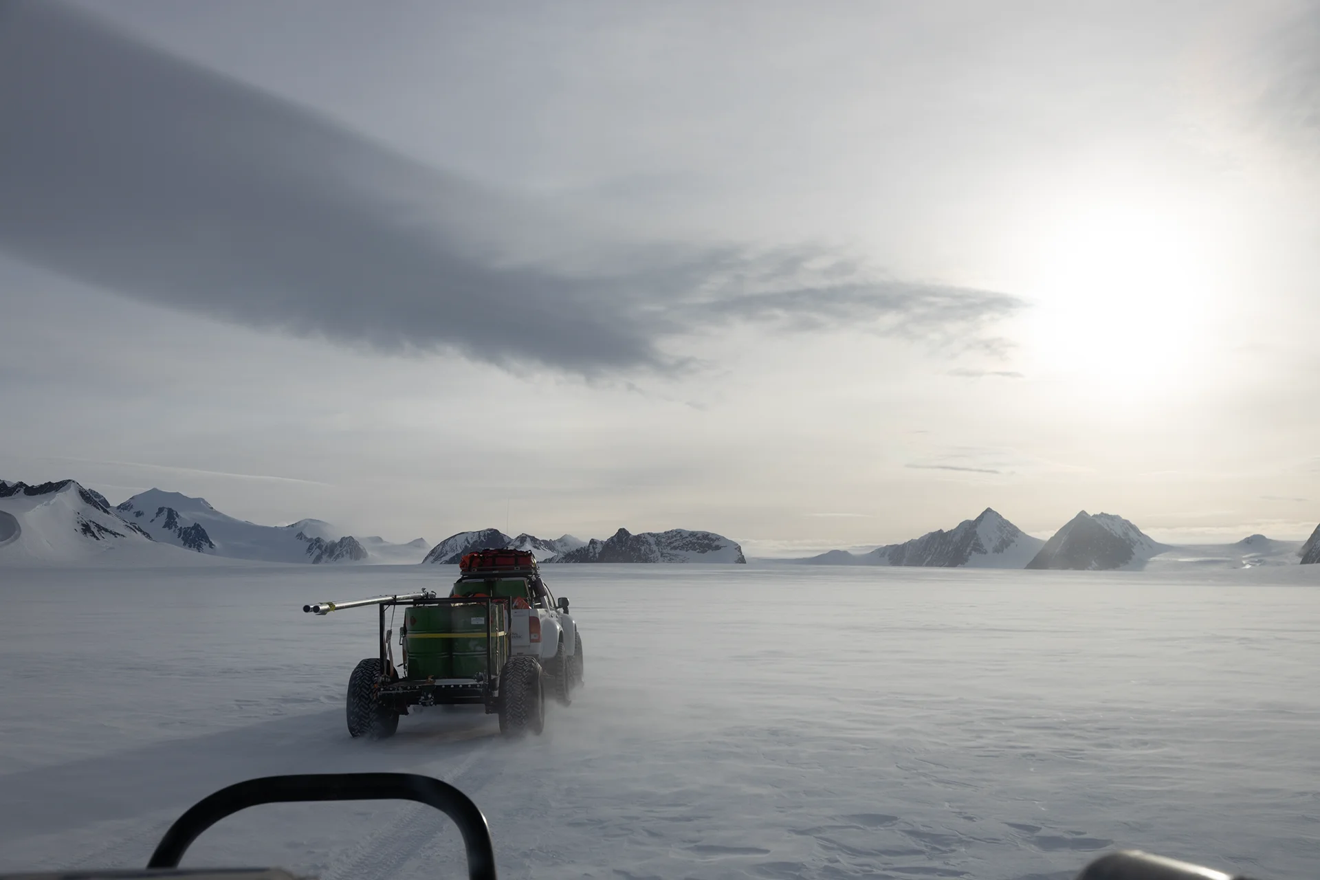 Truck towing equipment across a snowy landscape with distant mountains under a bright sky.