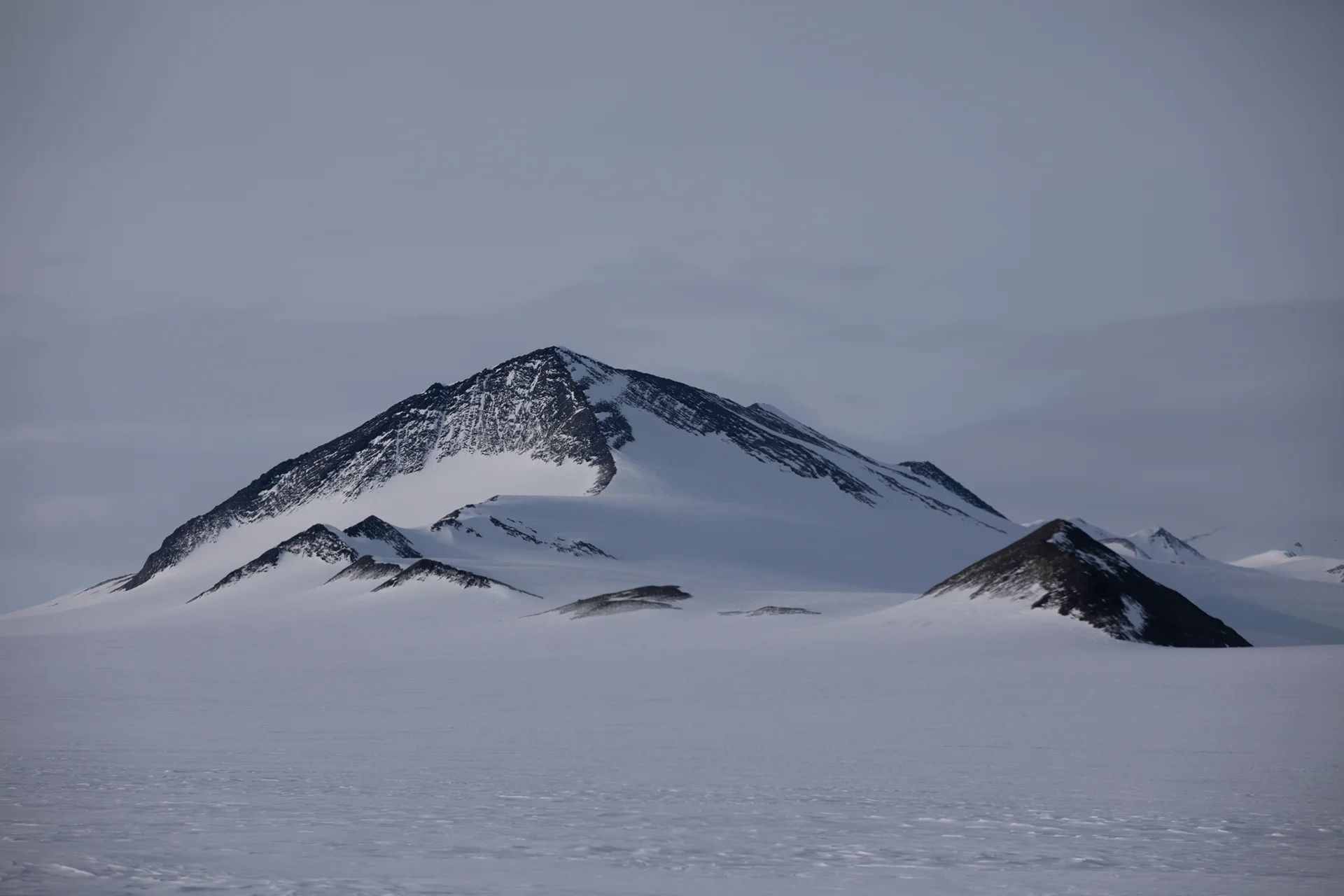 Snow-covered mountain peaks with dark rocky ridges under a gray cloudy sky.