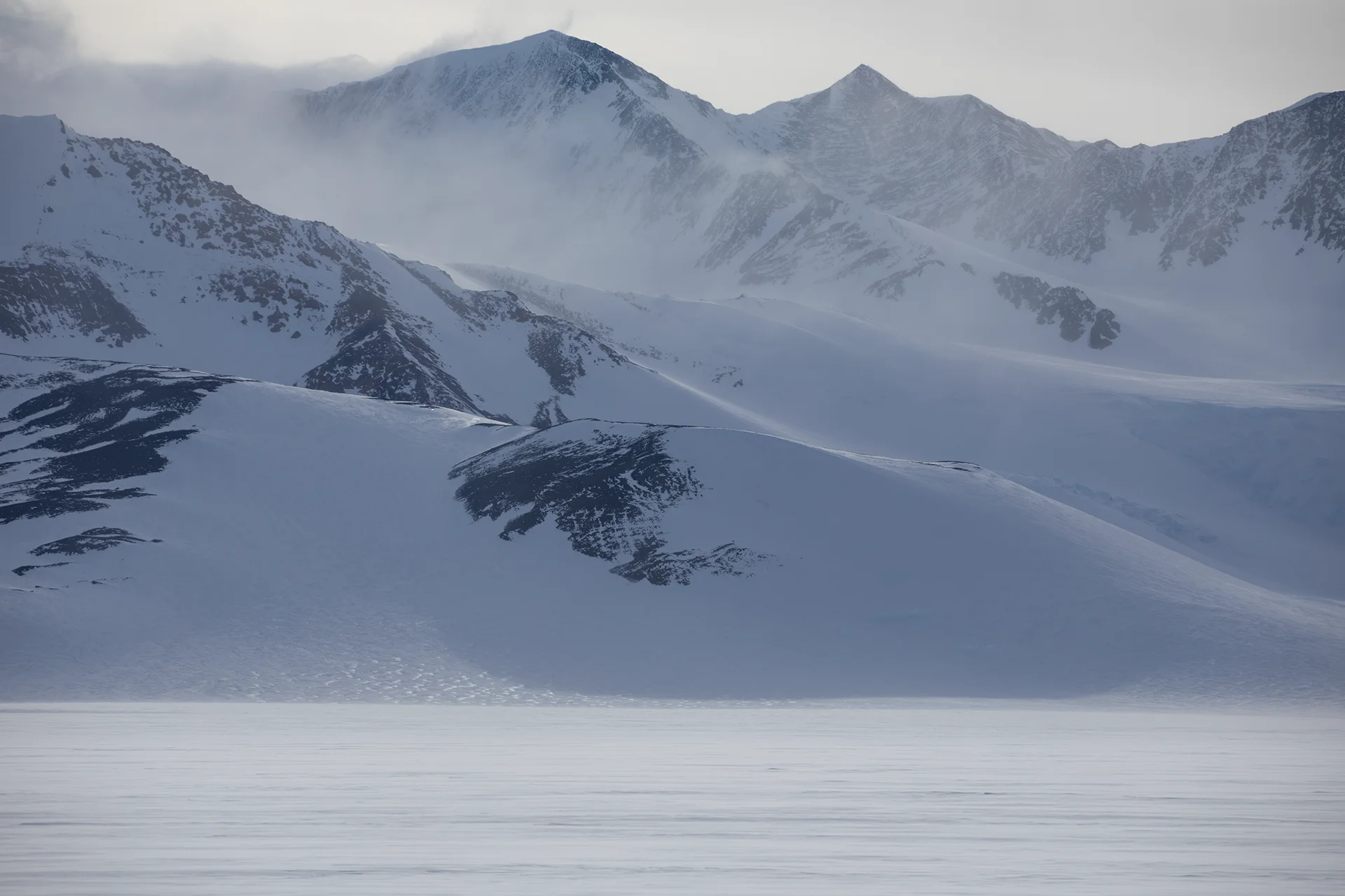Snow-covered mountain peaks and ridges under a cloudy sky with mist in a cold landscape.