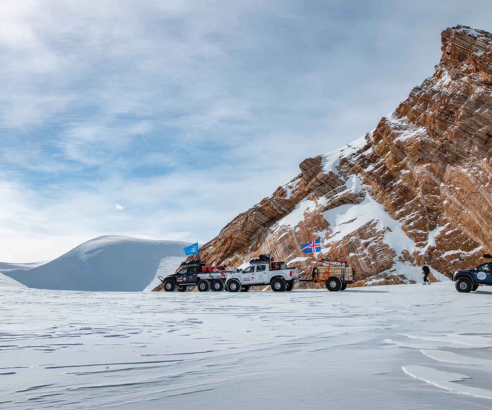 Four trucks parked on snowy terrain by a rocky, snow-dusted mountain under a cloudy blue sky.