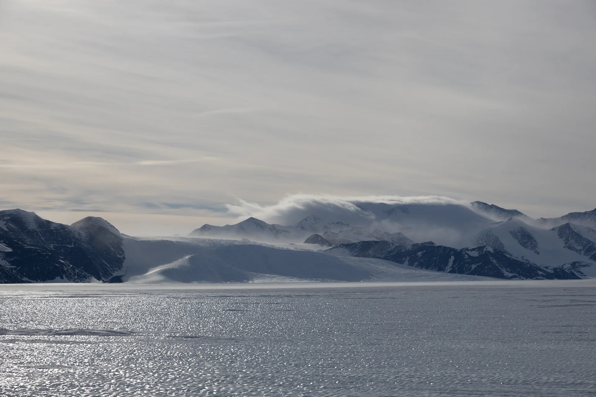 Snow-covered mountains with clouds and ice or frozen water reflecting light in foreground under overcast sky.