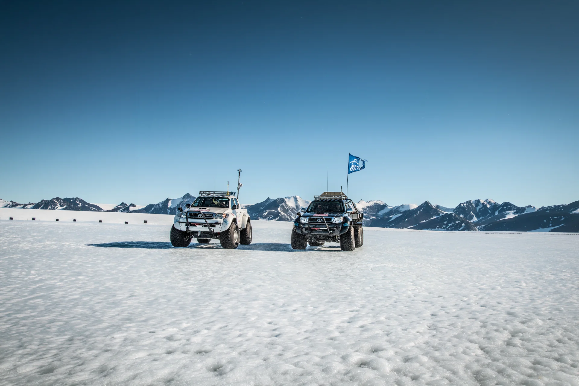Two off-road vehicles on snow with a clear sky and mountains in the background.