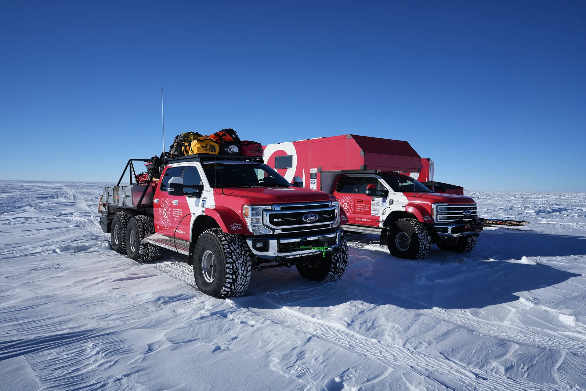 Two red and white off-road trucks with large tires parked on snow under a clear blue sky.