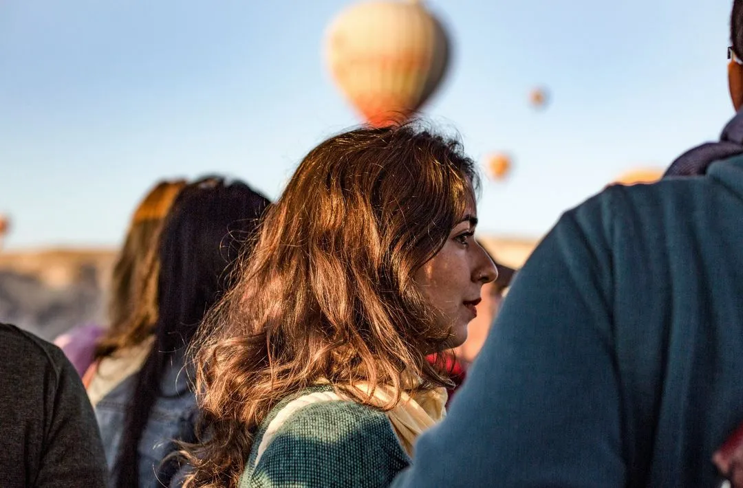 Profile of a woman with long brown hair, softly lit by the sun, among a crowd. Hot air balloons float in the clear blue sky above, conveying a sense of wonder.