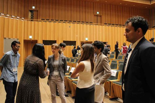 A group of diverse people in professional attire conversing and shaking hands inside a large auditorium with wooden walls and empty green seats.