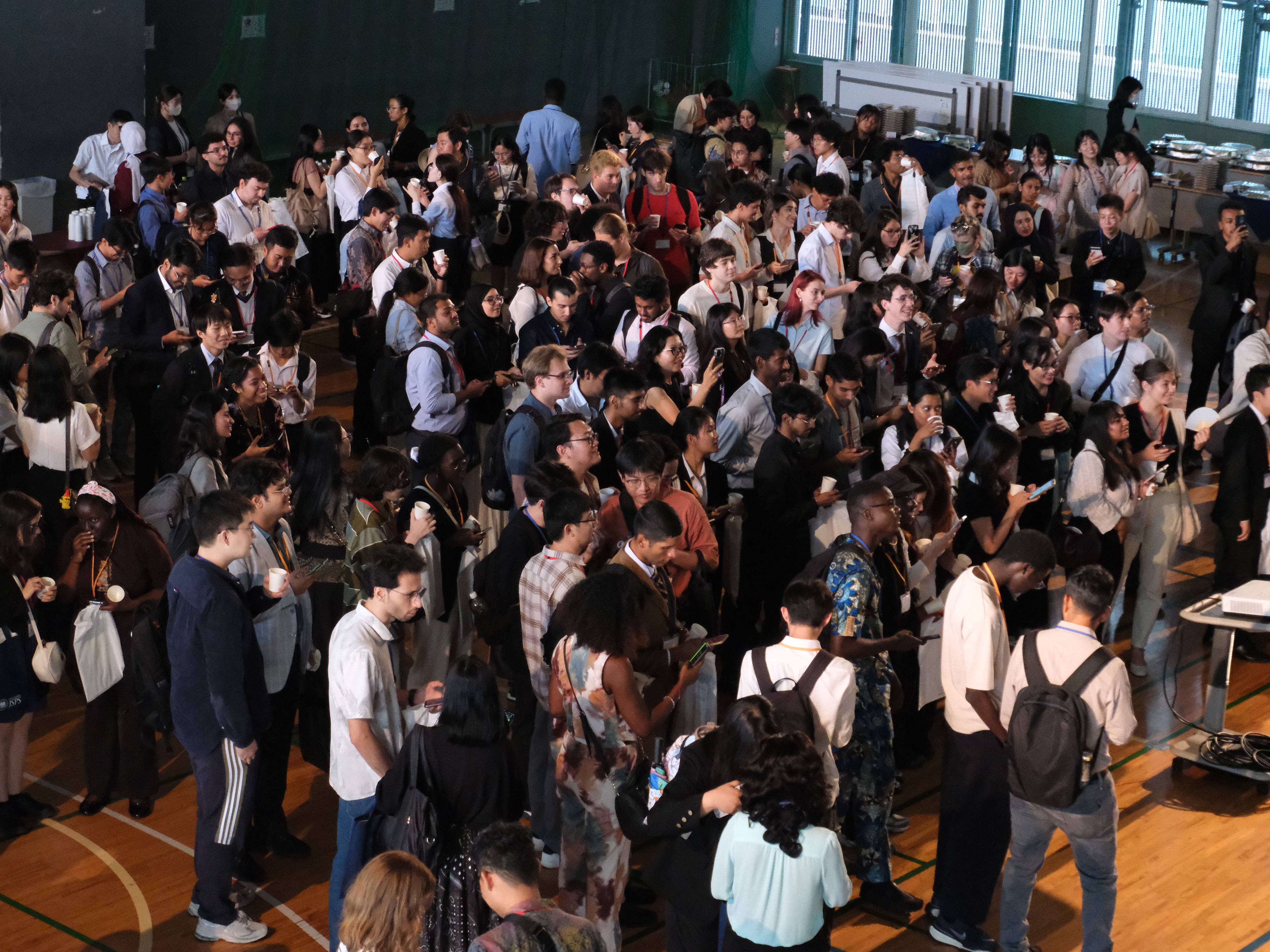 Large diverse group of people gathered indoors, many holding cups and some looking at their phones.