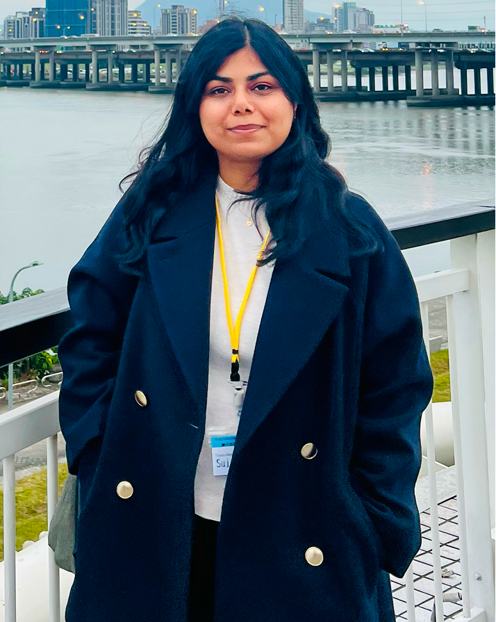 Woman with long dark hair wearing a navy blue coat and a white top, standing outdoors by a railing with a river and city buildings in the background.