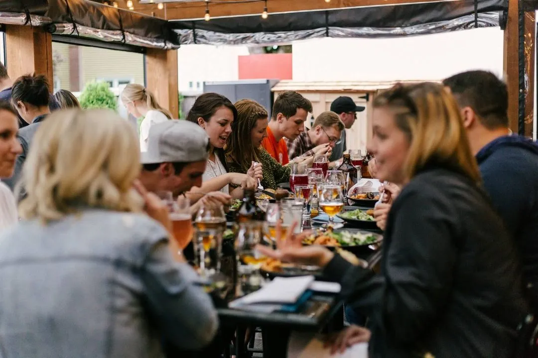 A group of people enjoying a lively outdoor dinner, sitting closely at a long table filled with plates of food and glasses. String lights create a warm atmosphere.
