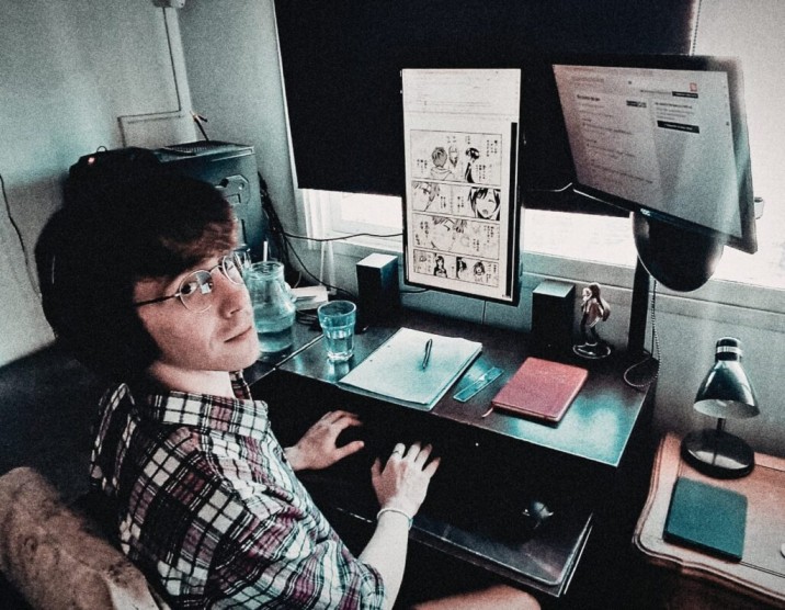 Man in plaid shirt speaking to a seated audience in a cozy, well-lit room with string lights. The atmosphere is engaging and focused.