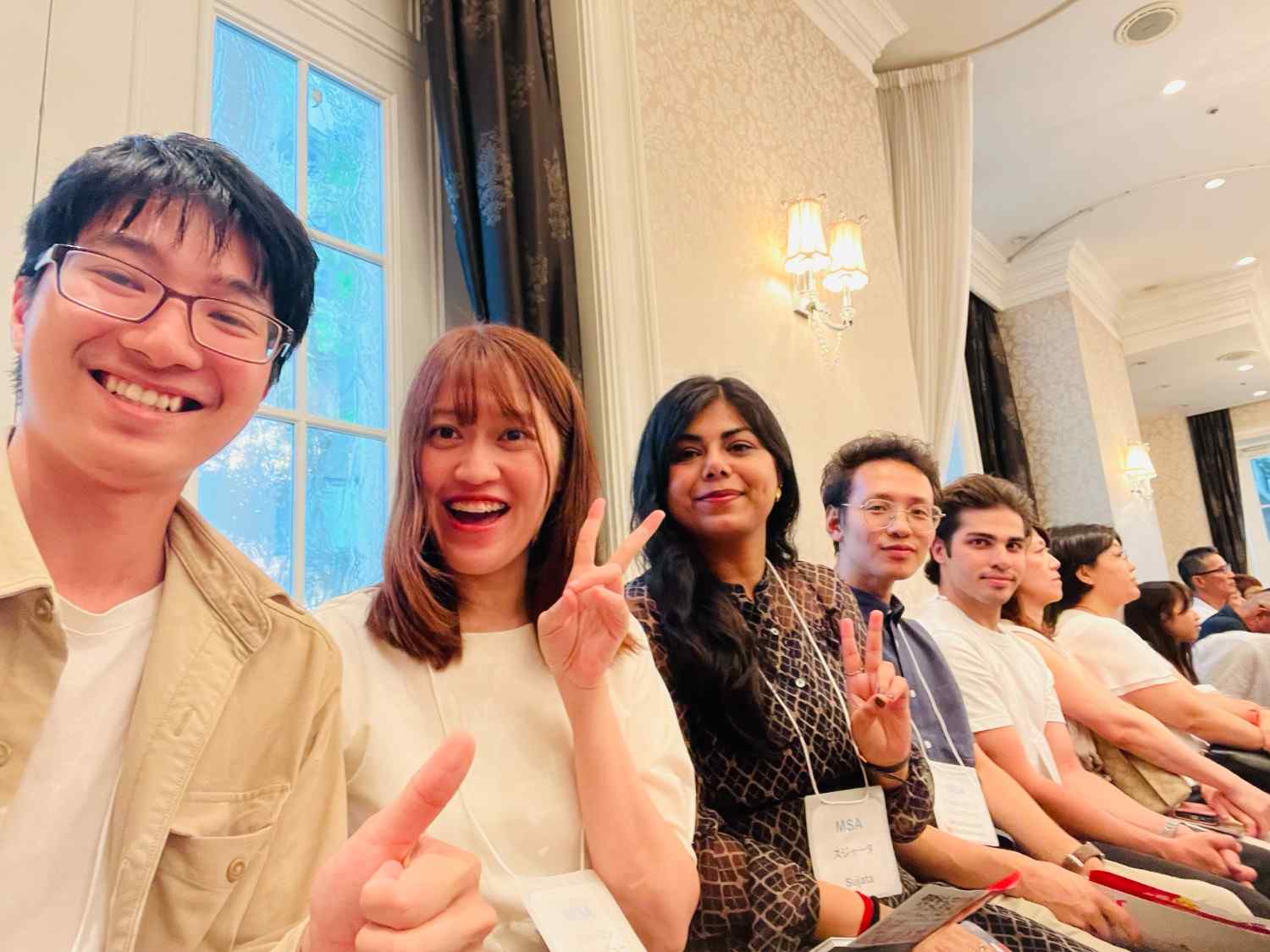 A lively crowd celebrates at a Pride parade under a sunny sky. People cheer and wave flags near a colorful float surrounded by lush green trees.