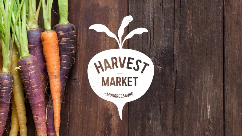 Colorful carrots with green tops on a wooden background beside a white logo reading 'Harvest Market Moorreesburg'.